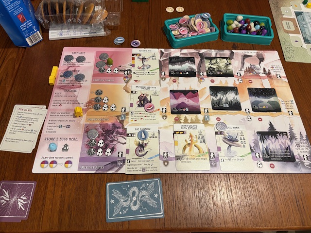 A board game board is out that has cards and tokens on it. There are additional cards, a box of small coin-sized cardboard tokens, and a box of small and similarly sized dragon eggs. 

The cards have different dragons on them.